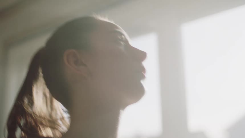 A woman in a white shirt leans forward in a softly lit room appearing to paint a wall near a window