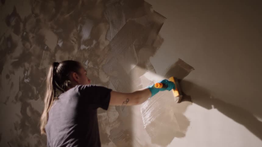 A woman with a ponytail and is using a paintbrush and blue gloves to paint a wall with brown paint