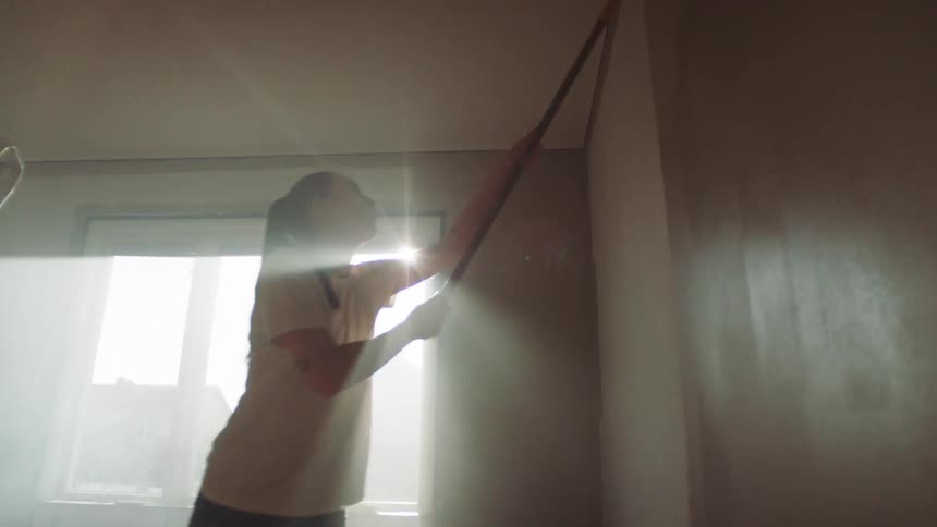 A young lady uses a paint roller to paint her room wall at midday