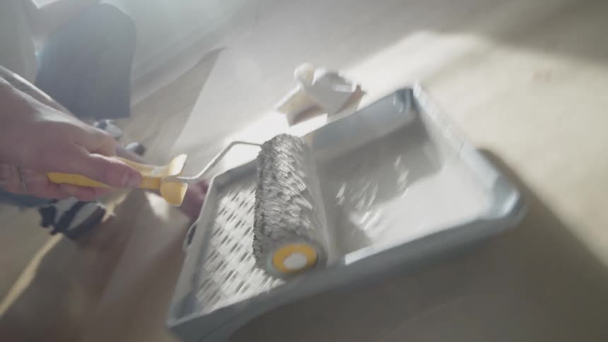 A person rolls a paint roller over a tray of paint in a sunlit room ready to begin painting a wall