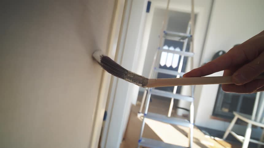 A persons hand holding a brush that is passing over the surface of a white wall to paint it and then placing it in a bucket