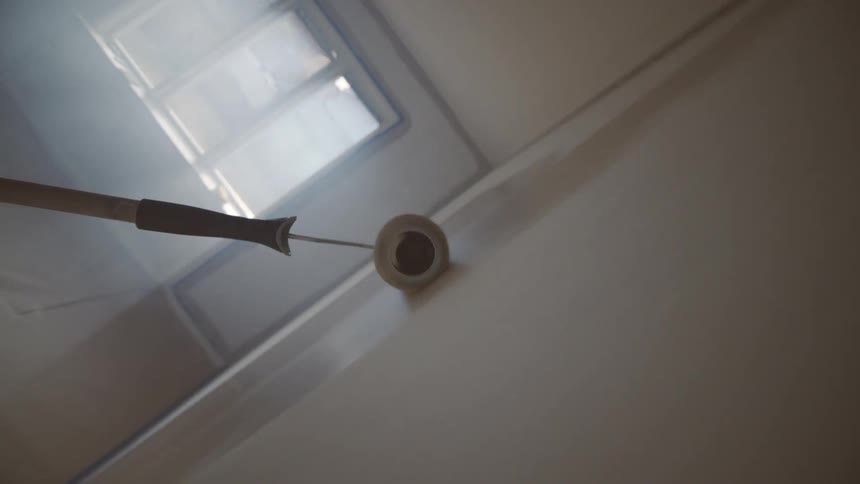 A paint roller spreads paint up and down on a building wall in a room at midday