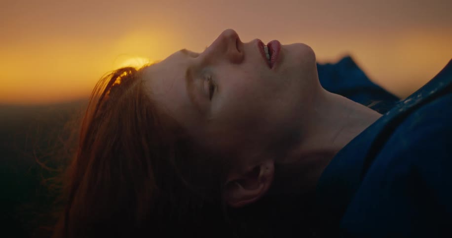 The face of a redhaired woman who is lying with her mouth partially open and behind her the sun shining brightly orange in the sky