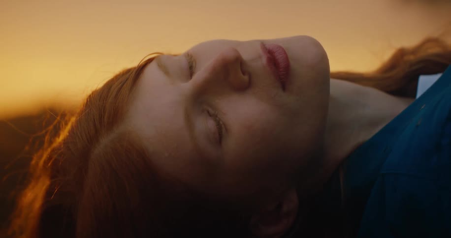 A woman with red hair wearing a blue dress lies outdoors as the orange glow of the setting sun casts a warm light over her