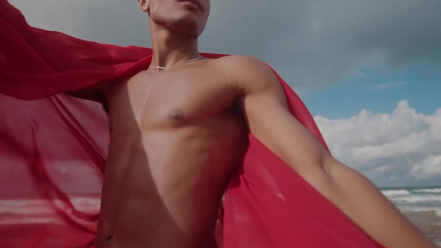 A man stands with a red piece of fabric draped around him while he stands on the beach