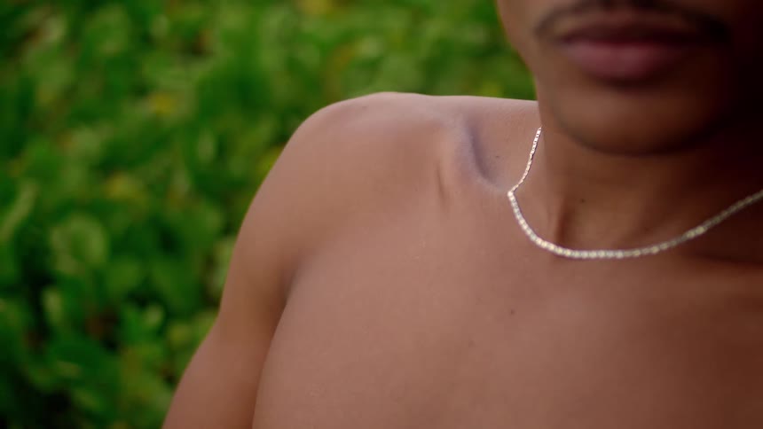 A shirtless man wearing a silver necklace sits beside a lawn around midday
