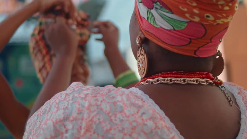 Two women adjust colorful headwraps one wearing a lace top and large earrings