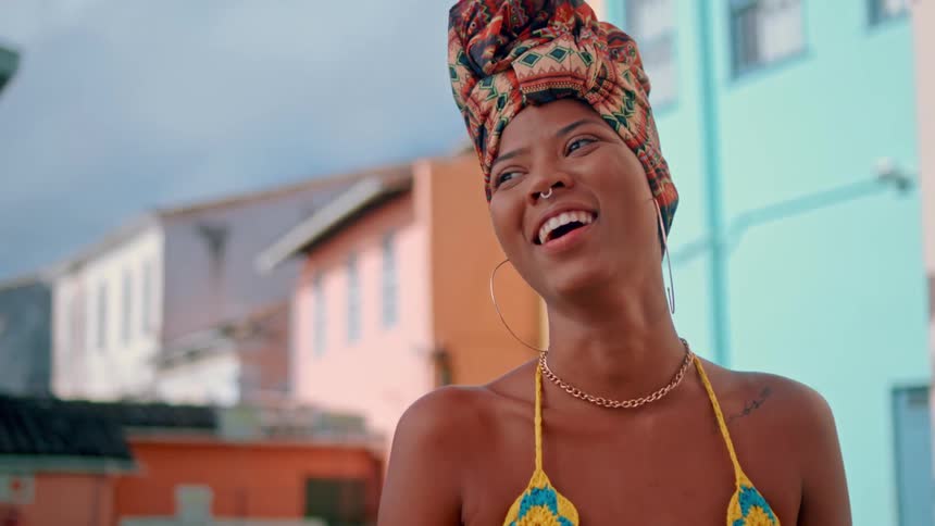 A smiling woman in colorful attire stands in front of vibrant buildings outdoors