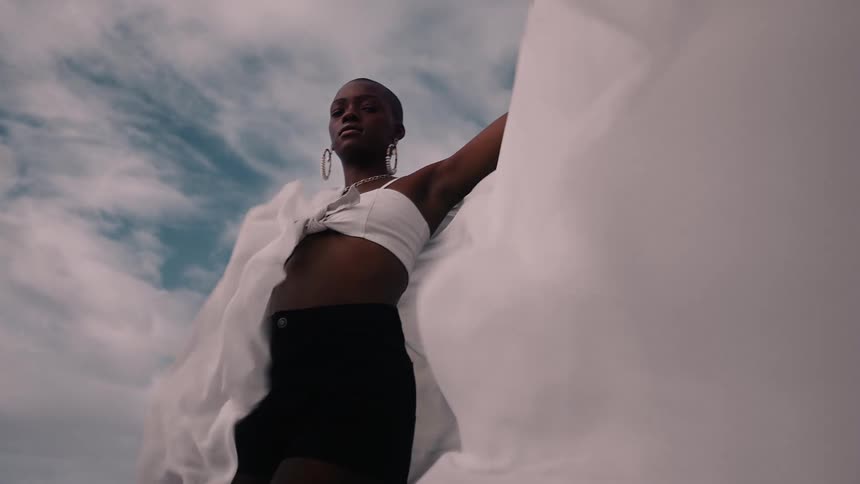 A confident woman poses dramatically with flowing fabric against a cloudy sky