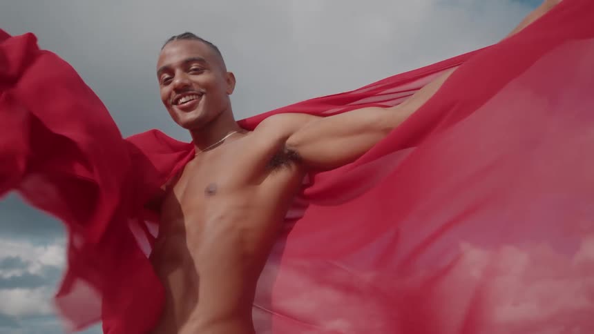 A joyful shirtless man poses outdoors holding a flowing red fabric against a bright sky