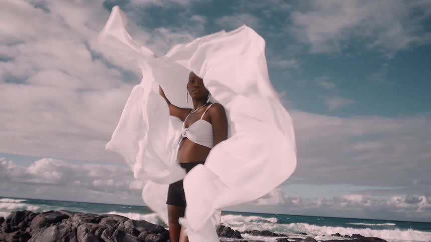A woman models on coastal rocks holding flowing white fabric as it billows in the wind