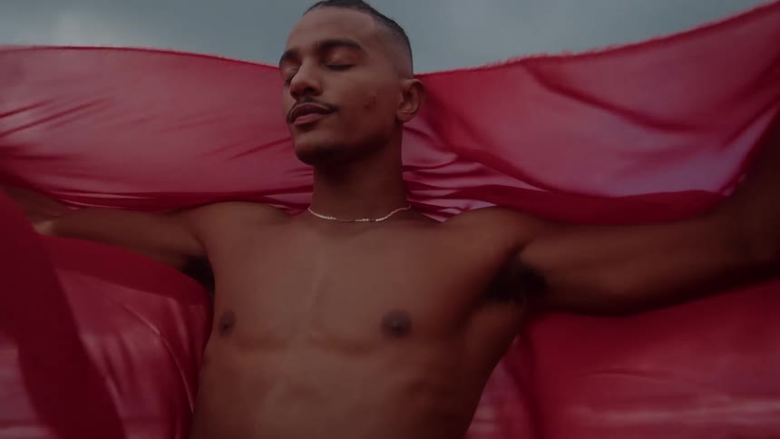 A young shirtless black man stands on the beach holding a red cape as the midday wind blows it behind him