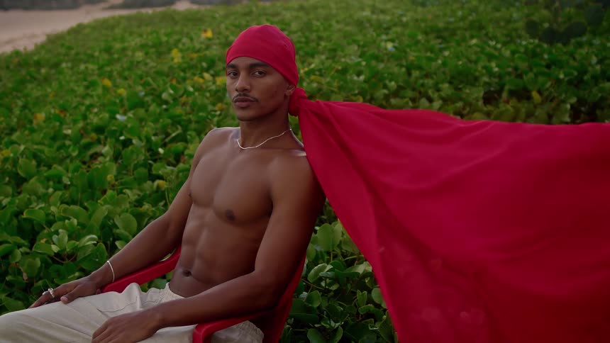 A young shirtless man is sitting in an environment with green vegetation behind him and wearing a huge red bandana that i was flying with the wind
