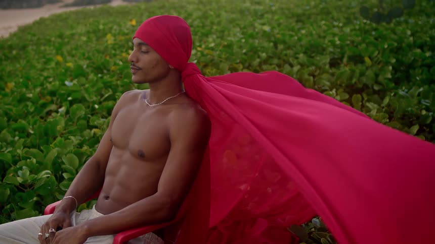 A young black man shirtless ties a red scarf on his head while sitting beside a garden as the wind blows the scarf at midday