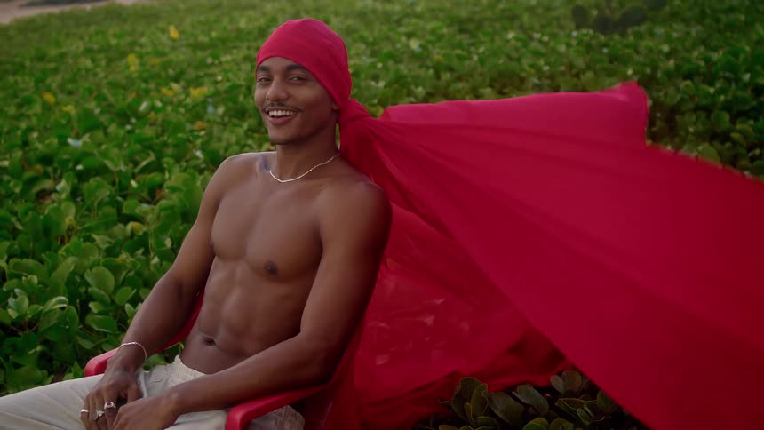 A man sits on a lawn wearing a red cape over his head as the wind blows the cape around him around midday