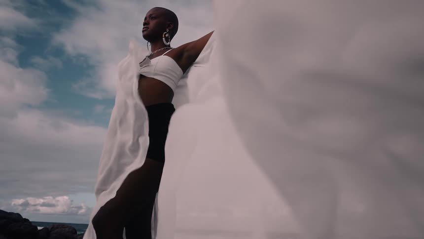 A woman poses dramatically on coastal rocks with flowing fabric billowing in the wind