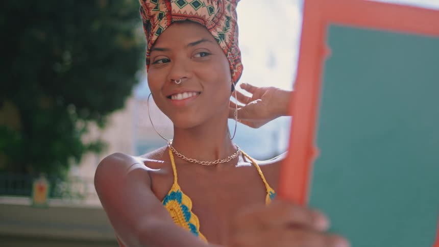 A woman wearing a colorful headwrap and a yellow patterned top holds up an orangeframed mirror while standing outdoors