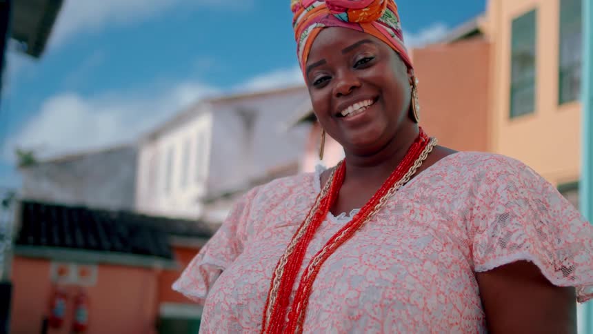 A young woman wearing a gown and several necklaces smiles while standing outside at midday