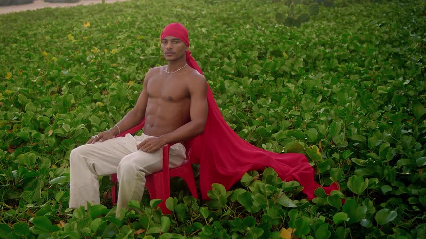 Man sits in a field of plants wearing a long fabric headdress that matches his chair in color