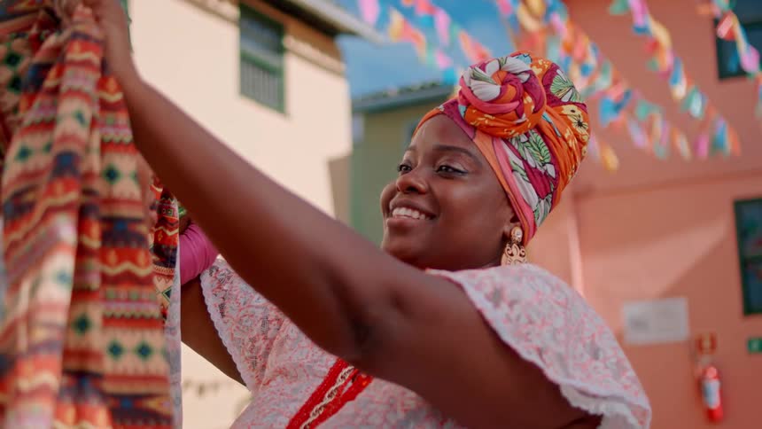A woman adjusts the vibrant patterned headscarf of another in an outdoor area decorated with colorful string flags