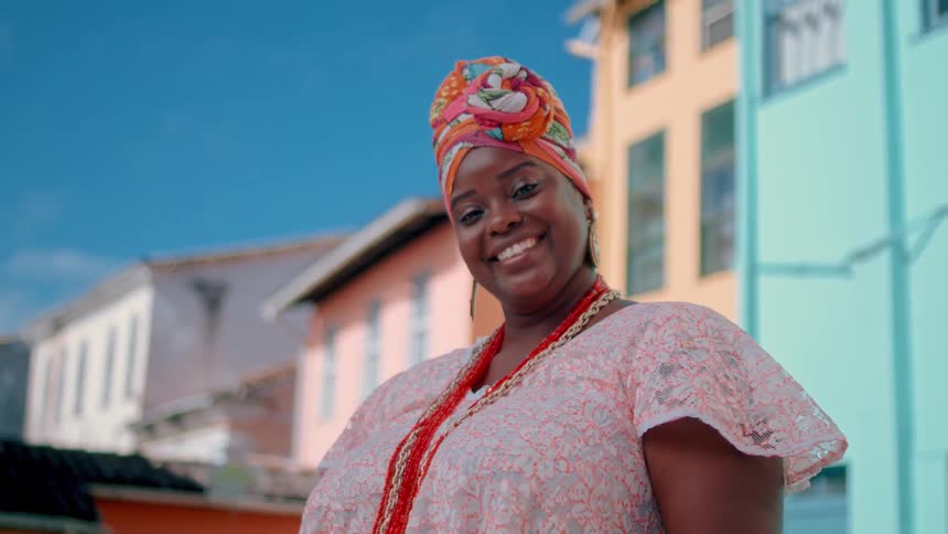 A smiling woman adorned in a colorful headwrap and multiple orange beaded necklaces stands outdoors with brightly painted buildings under a clear blue sky in the background