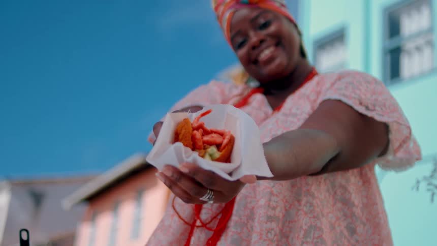 A young black woman wearing a gown hair tie and several beads around her neck stands outside a building at midday holding a piece of seafood