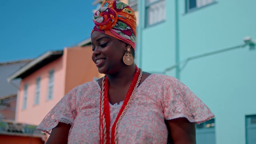 A young woman wearing a headtie a gown and a necklace stands outdoors smiling