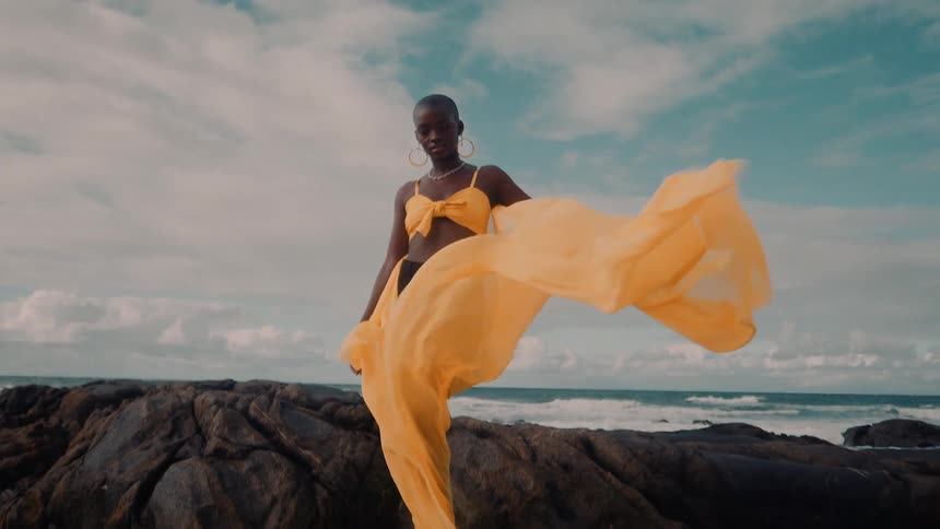 A woman standing on the rocks of a beach holding the skirt of her yellow dress which is flying in the wind
