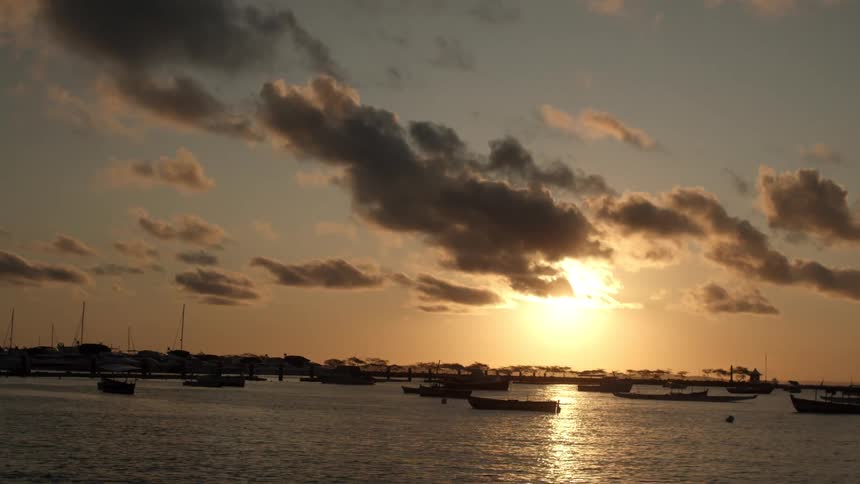 The golden light of a setting sun shimmers on the water of a calm bay silhouetting the many small boats floating in the harbor