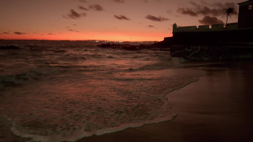Gentle waves wash onto a sandy beach at sunset with the silhouette of a coastal fort against the orange sky and distant city lights