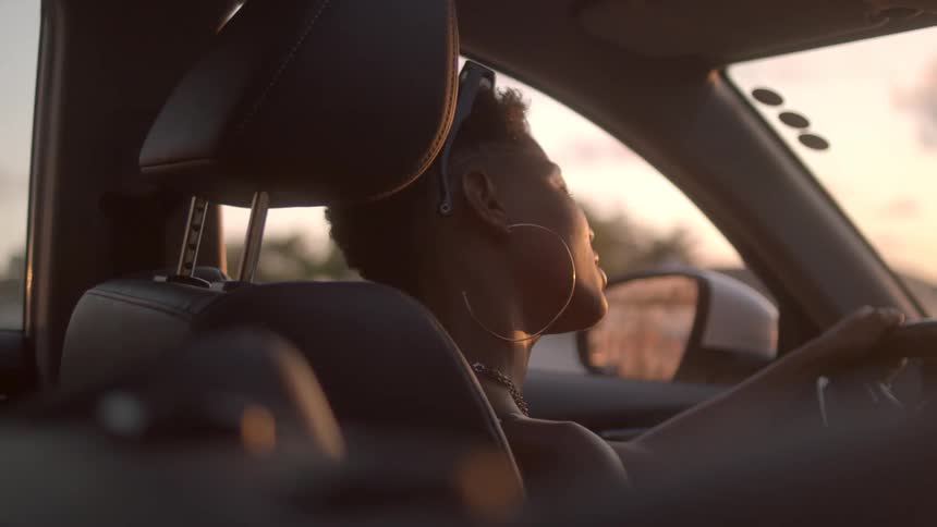 A young black woman drives a car at midday with a smile on her face