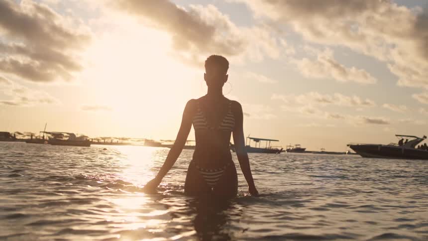A silhouetted woman in a bikini emerges from the ocean walking slowly towards the camera as the golden sun sets behind her