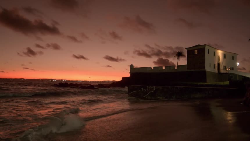 Waves crash against a coastal building as the sky glows orange at sunset
