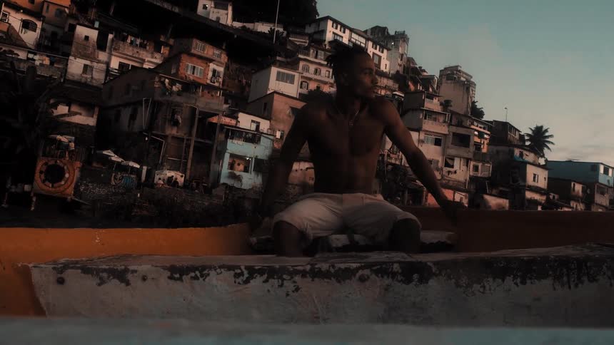 Shirtless man sits gazing beside hillside housing