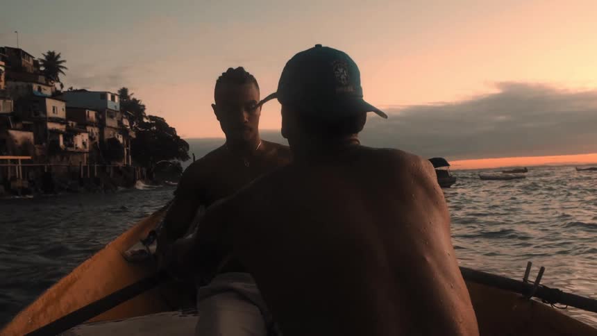 Two people row a small boat across calm ocean waters near a coastal village under a vivid orange sunset