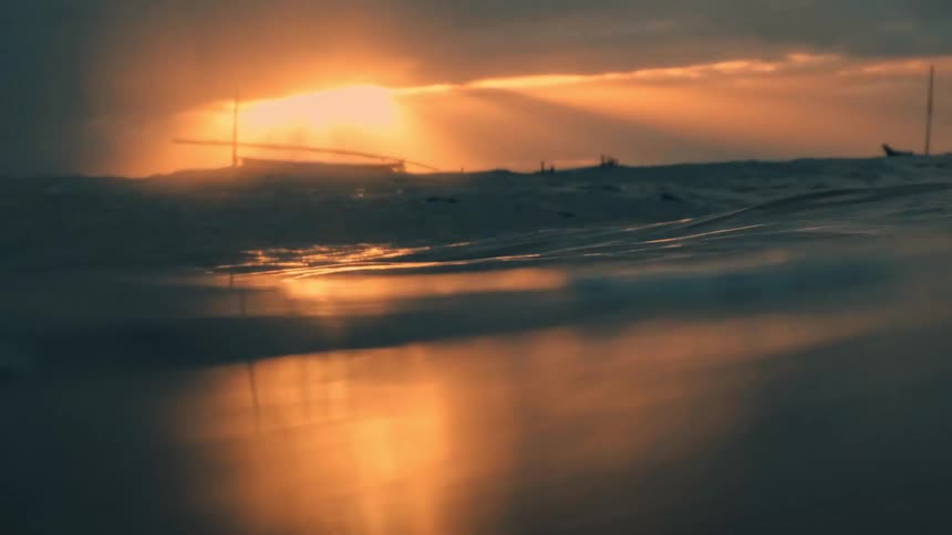 The camera moves through rough ocean waves partially submerging before revealing a boat silhouetted against a dramatic sunrise with sun rays piercing through the clouds