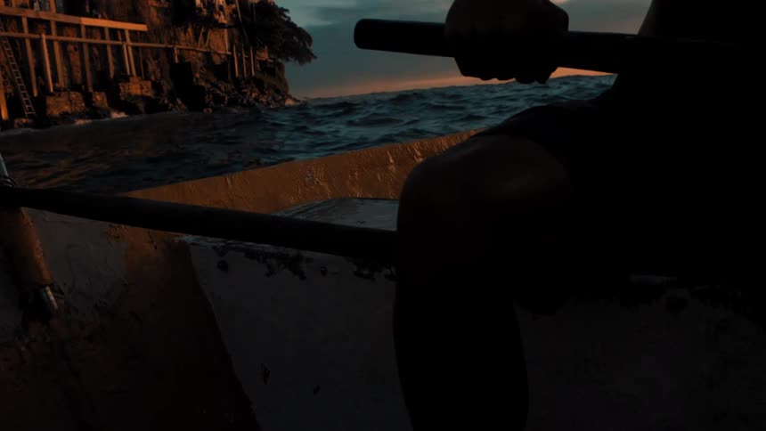Man sits in a rowboat near coastal buildings while red tints the horizon