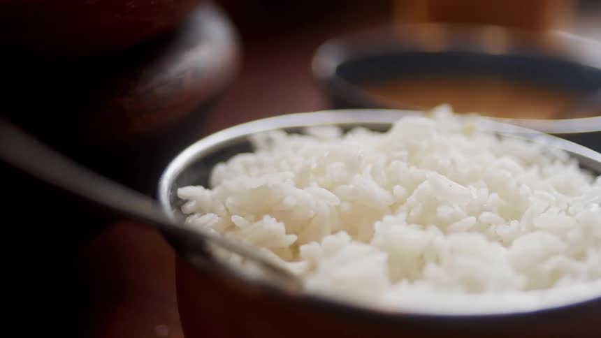 A pot full of white rice with a metal utensil on the edge and a black bowl with soup behind it