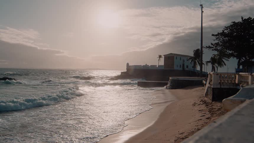 The sun shines brightly over a coastline where waves crash onto a sandy beach next to a stone wall and a distant historic building