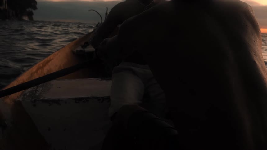Two shirtless men are in a canoe at sundown with one paddling while the other watches