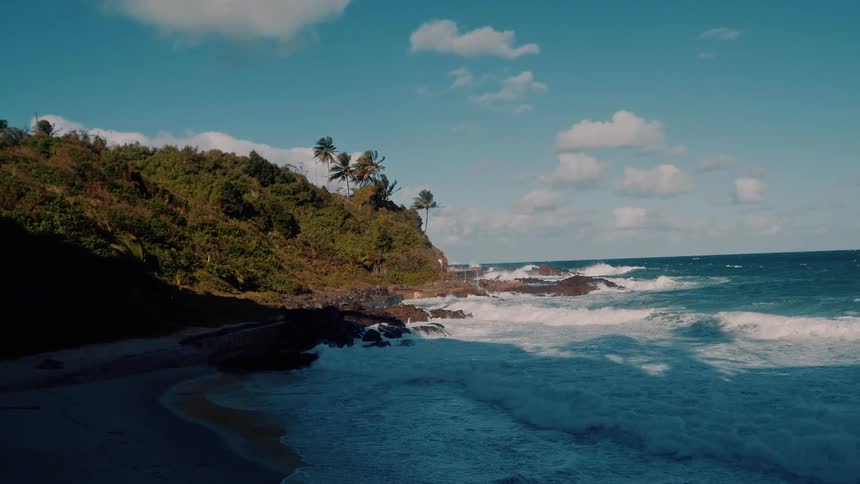 An aerial shot reveals waves crashing on a tropical shoreline with a coastal road running alongside a green hill under a blue sky