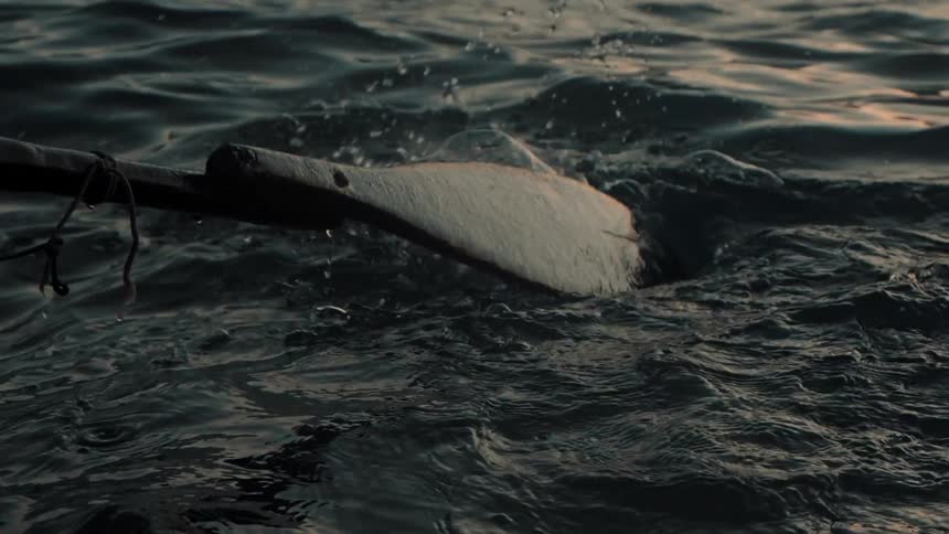 From the perspective of a bobbing boat a weathered wooden oar repeatedly dips and pulls through dark water at dusk