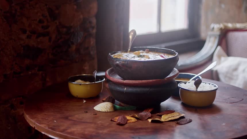 A rustic table is set with steaming bowls of food and scattered dried ingredients in a cozy room