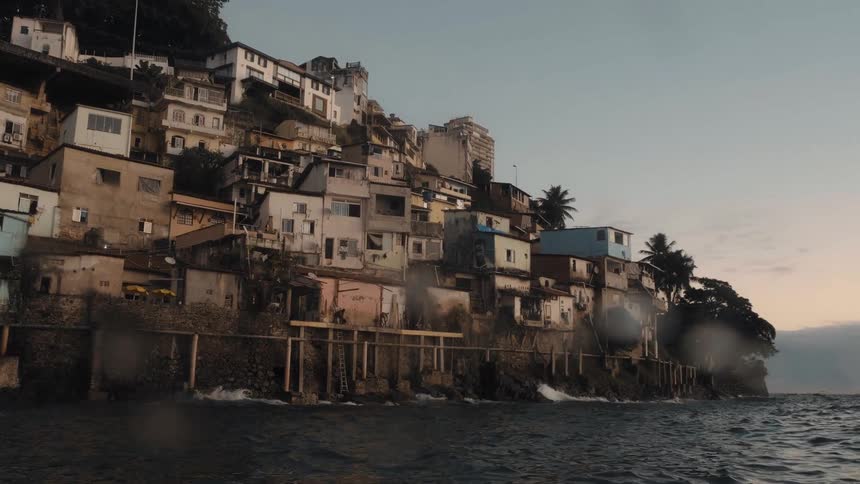 Viewed from a moving boat a dense favela community is packed onto a steep hillside that meets the oceans edge at sunset