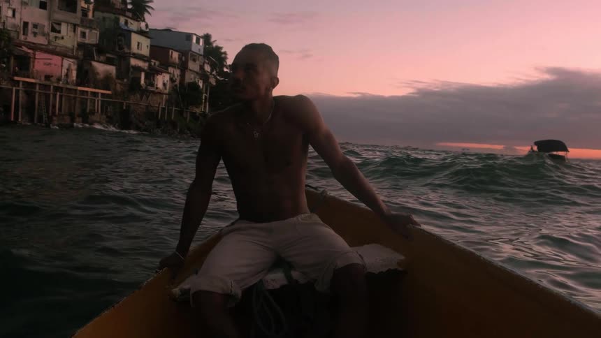 A shirtless man sits in a small boat at dusk looking toward the shoreline
