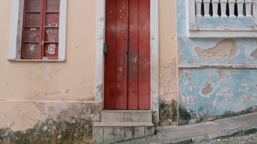 A tall red door stands between two weathered colorful old buildings on a sloped street showing peeling paint and signs of age