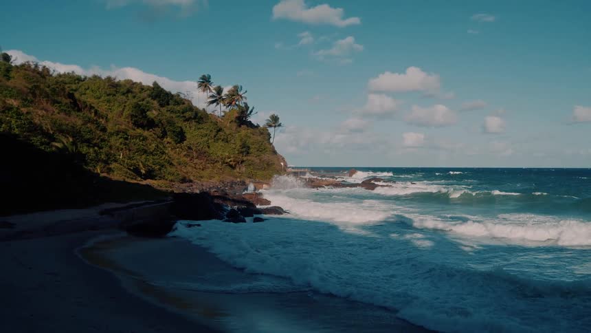 A seaside road runs along a tropical beach where waves crash against a rocky palmtopped cliff under a partly cloudy sky