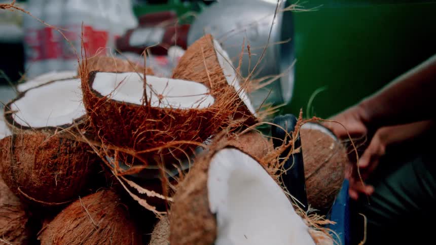 A person grates fresh coconut at a streetside stall surrounded by halved coconuts