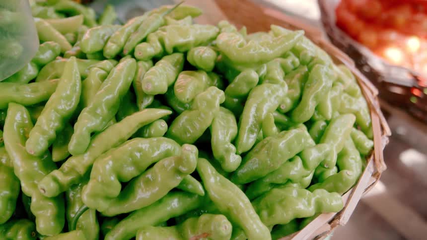 A closeup shot captures a large pile of fresh light green chili peppers filling a woven basket at a market