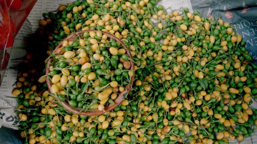 A large pile of fresh green and yellow chili peppers sits on newspaper at a market with a small basket full of them resting in the center